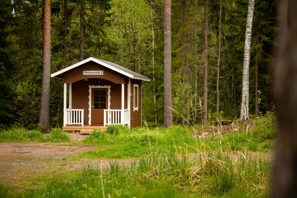 Portinvartijat 1, 2 ja 3 Pieni, jopa piparkakkutalomainen Portinvartija 3 -mökki kuvattuna kauempaa. Mökki seisoo metsän laidalla.