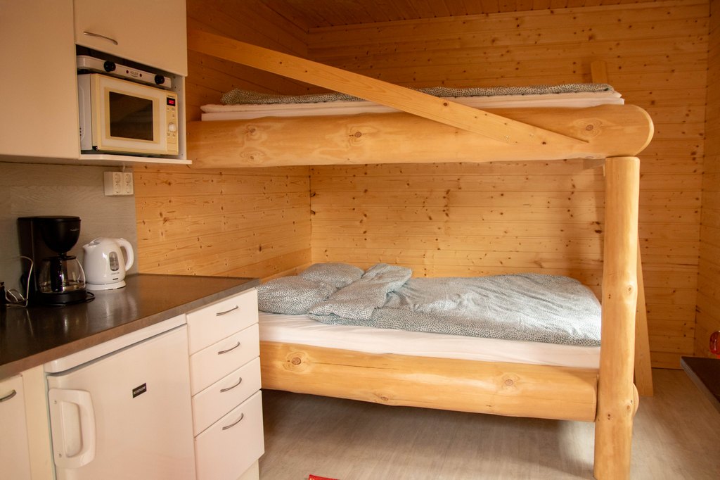 Portinvartijat 1, 2 ja 3 Kelopuista rakennettu kerrossänky, jossa sinertävät lakanat. Etualalla myös pieni keittiösaareke.